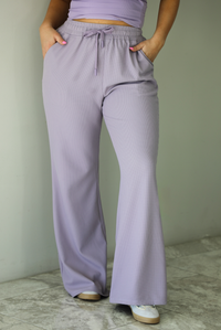 Woman wearing a two piece lavender set featuring a strapless, fitted tube top, and high waisted, flared hem pants. The photo is taken indoors against a white wall and shows a closeup of the pants, which are high waisted with an elastic and drawstring waistline, with functional pockets on either side, has a repeating squared textured design, and a flared hem.