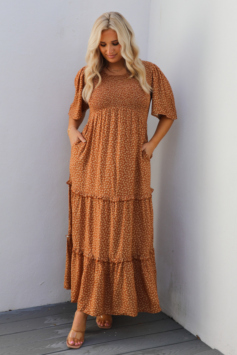 A woman model outdoors wearing a rust dress with a cream floral print pattern throughout. It has flowy short sleeves, a smocked bodice, and a flowy maxi length skirt with functional pockets on either side. 