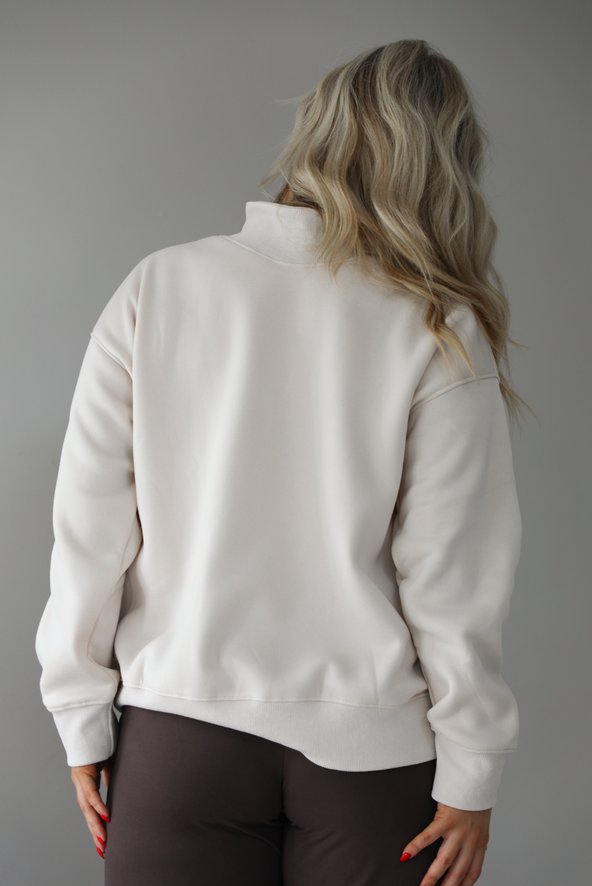 Woman wearing a beige high-neck, oversized sweaters and high waisted brown pants. The photo is taken indoors against a white wall with white marbled flooring. The photo shows a closeup of the back of the sweater.