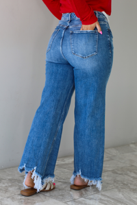 Woman wearing a pair of medium wash denim that are high waisted, have functional button and zipper closures, functional front and back pockets, a straight leg design, and a raw, distressed hem. The photo shows the back of the denim, with the models hand in the back right pocket. The photo is taken indoors against a white wall with white marble flooring. 