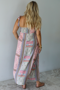 A woman wearing a thick strapped maxi dress with a patchwork print of pink gingham, beige paisley, and blue patterns, standing indoors against a white wall with white marble flooring. The photo shows the back of the dress. 