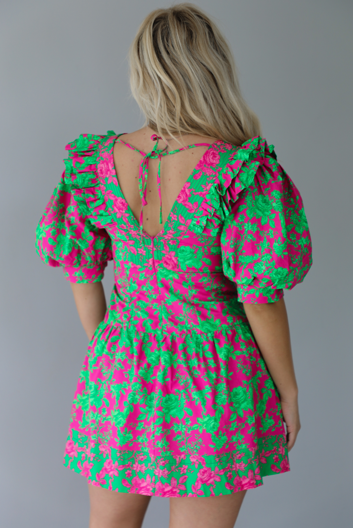 Woman wearing a fuchsia and green dress that has puffed sleeves, a v-neckline, fitted bodice, and flared skirt with pleated details indoors against a white wall. The photo shows the back of the dress, which has a zipper and tie closure. 