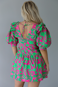 Woman wearing a fuchsia and green dress that has puffed sleeves, a v-neckline, fitted bodice, and flared skirt with pleated details indoors against a white wall. The photo shows the back of the dress, which has a zipper and tie closure. 