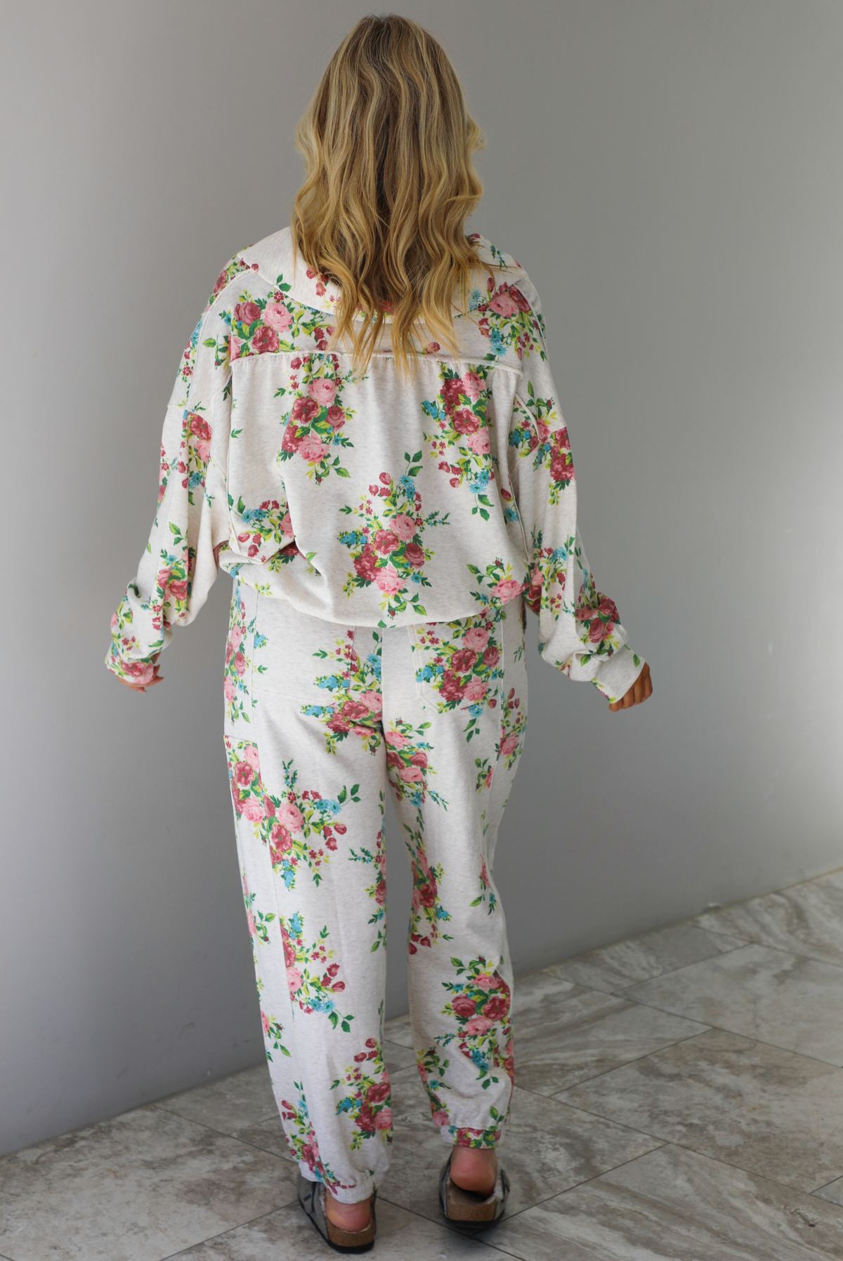Woman wearing an oatmeal colored and multi-colored floral print two piece set featuring a collared long sleeved crop top and high waisted pants. The photo shows the back of the set.  The photo is taken indoors against a white wall with white marble flooring. 