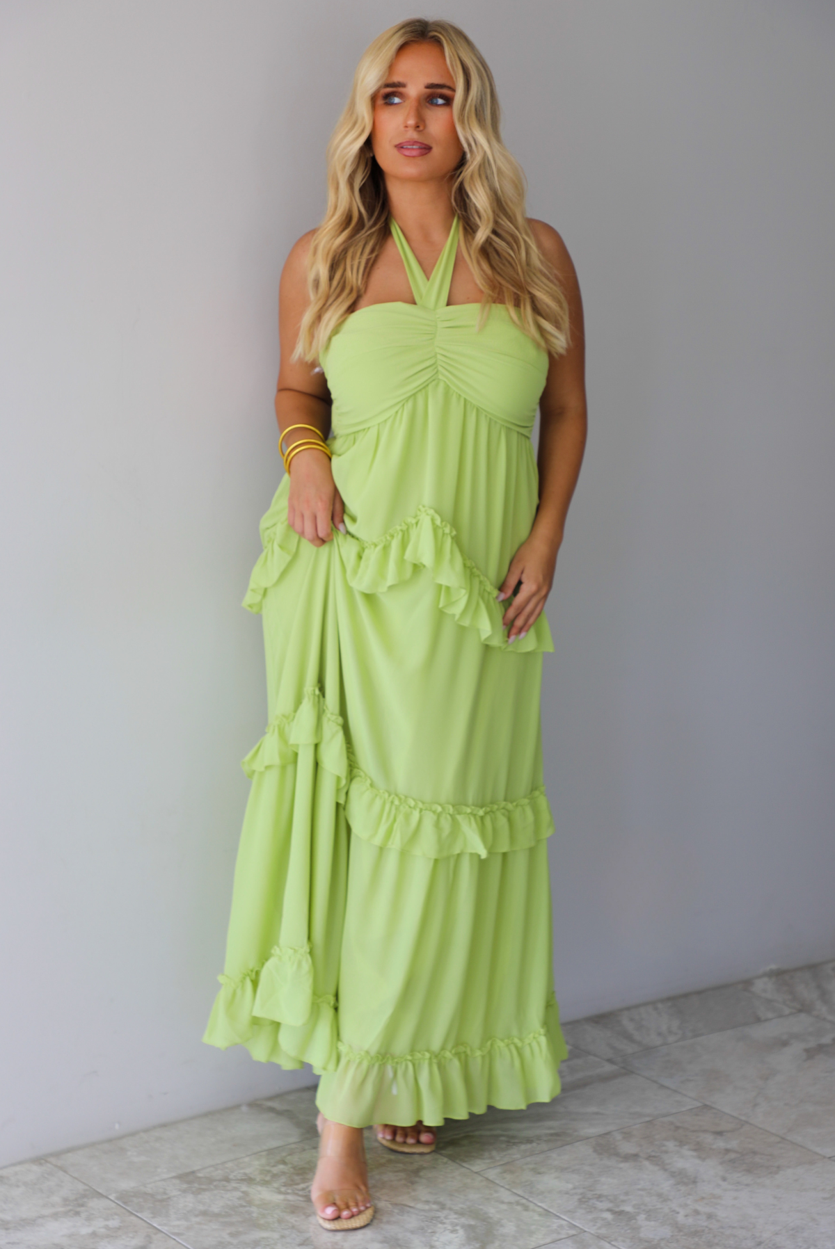 A woman modeling a lime green halter-neck maxi dress with tiered ruffle details, standing indoors against a white wall with white marble flooring. 