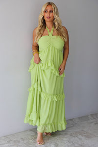A woman modeling a lime green halter-neck maxi dress with tiered ruffle details, standing indoors against a white wall with white marble flooring. 