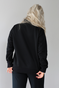 Woman wearing a black high-neck, oversized sweater and high waisted black pants. The photo shows the back of the sweater. The photo is taken indoors against a white wall with white marbled flooring. 