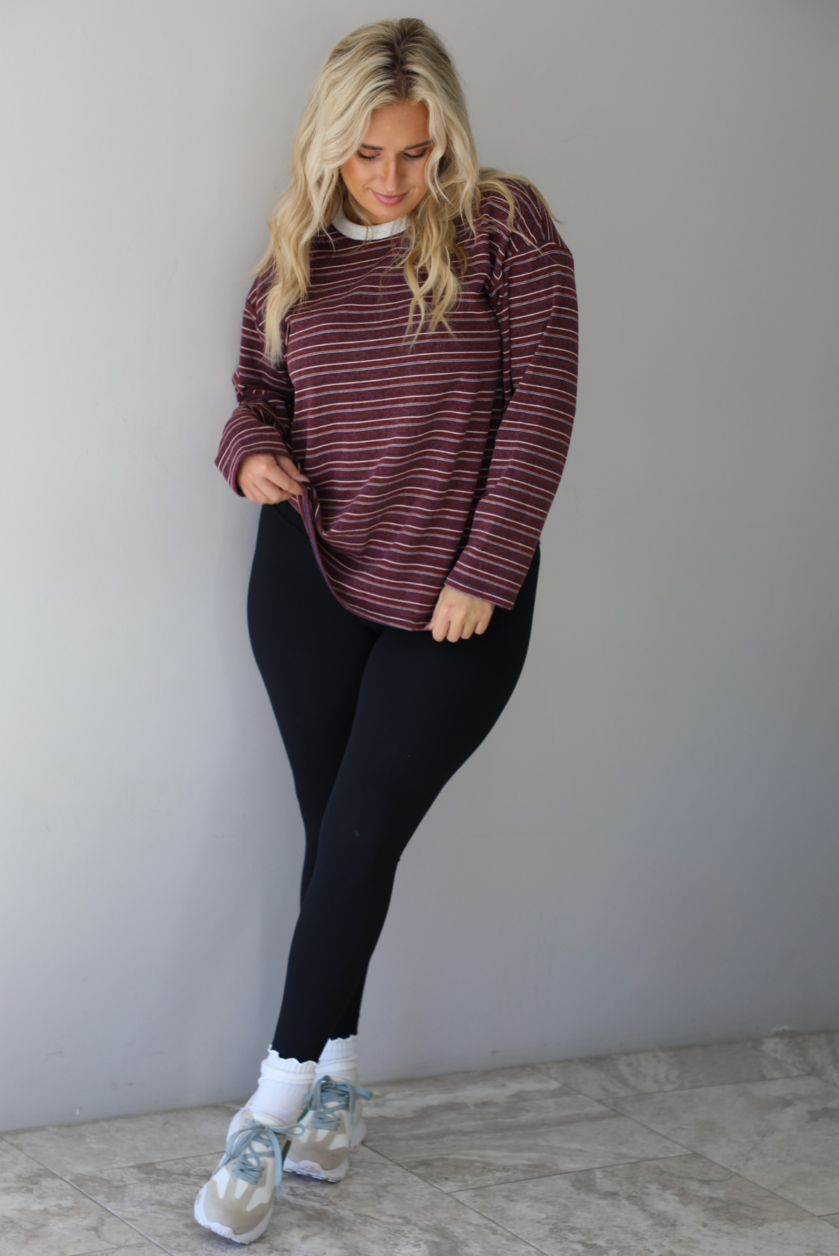 A woman wearing a burgundy striped long sleeved top paired with black leggings, high ankle socks, and sneakers, standing indoors against a white wall with white marble flooring. 