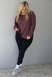 A woman wearing a burgundy striped long sleeved top paired with black leggings, high ankle socks, and sneakers, standing indoors against a white wall with white marble flooring. 