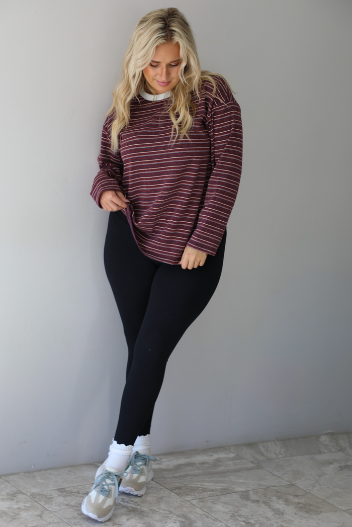 A woman wearing a burgundy striped long sleeved top paired with black leggings, high ankle socks, and sneakers, standing indoors against a white wall with white marble flooring. 