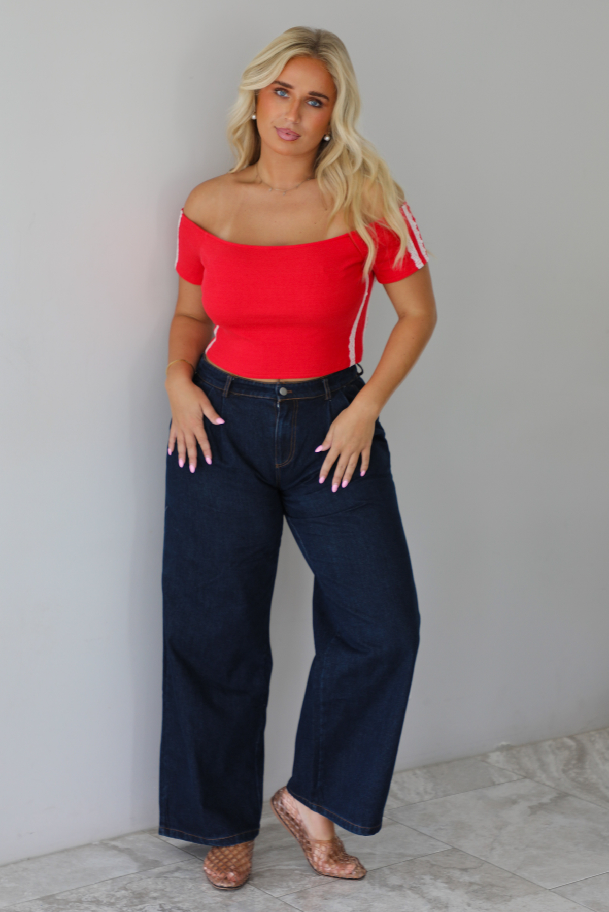 A blonde woman modeling a bright red off-the-shoulder short-sleeve top with white stripes on the sleeves, paired with dark wide-leg denim jeans, standing indoors against a white wall with white marble flooring. 