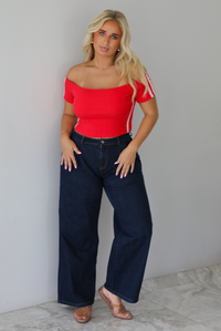 A blonde woman modeling a bright red off-the-shoulder short-sleeve top with white stripes on the sleeves, paired with dark wide-leg denim jeans, standing indoors against a white wall with white marble flooring. 