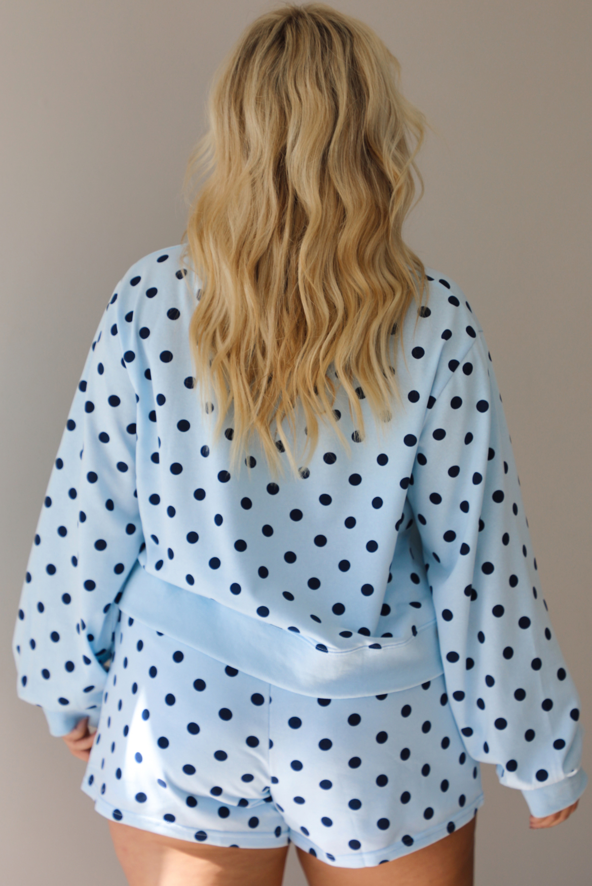 A woman shown from behind wearing a blue two piece set with a navy polka dot pattern throughout, featuring a sweatshirt and high waisted sweat-shorts, shown indoors against a white wall.