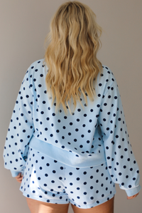 A woman shown from behind wearing a blue two piece set with a navy polka dot pattern throughout, featuring a sweatshirt and high waisted sweat-shorts, shown indoors against a white wall.