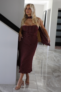 A woman wearing a strapless maroon midi dress with a white polka dot pattern with a matching scarf, standing inside a modern home with white marble flooring. 