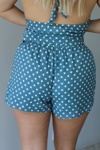 Woman wearing denim blue with white polka dot halter tie top and high waisted shorts indoors against a white wall. The photo shows a closeup of the back of the shorts. 