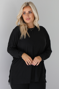 Woman wearing a black two piece set that features an oversized long sleeved top and fitted leggings. The photo shows a closeup of the top. The photo is taken indoors against a white wall.