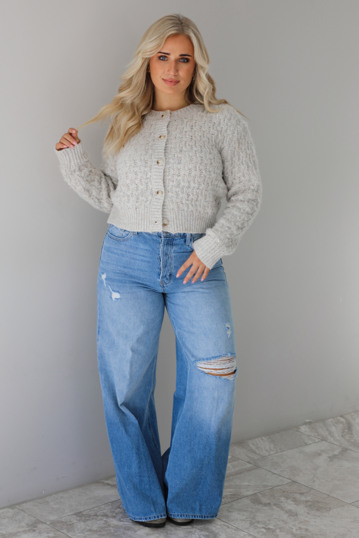 Woman wearing a light grey cardigan buttoned up with high waisted flared hem medium wash denim. The photo is taken indoors against a white wall with white marble flooring.
