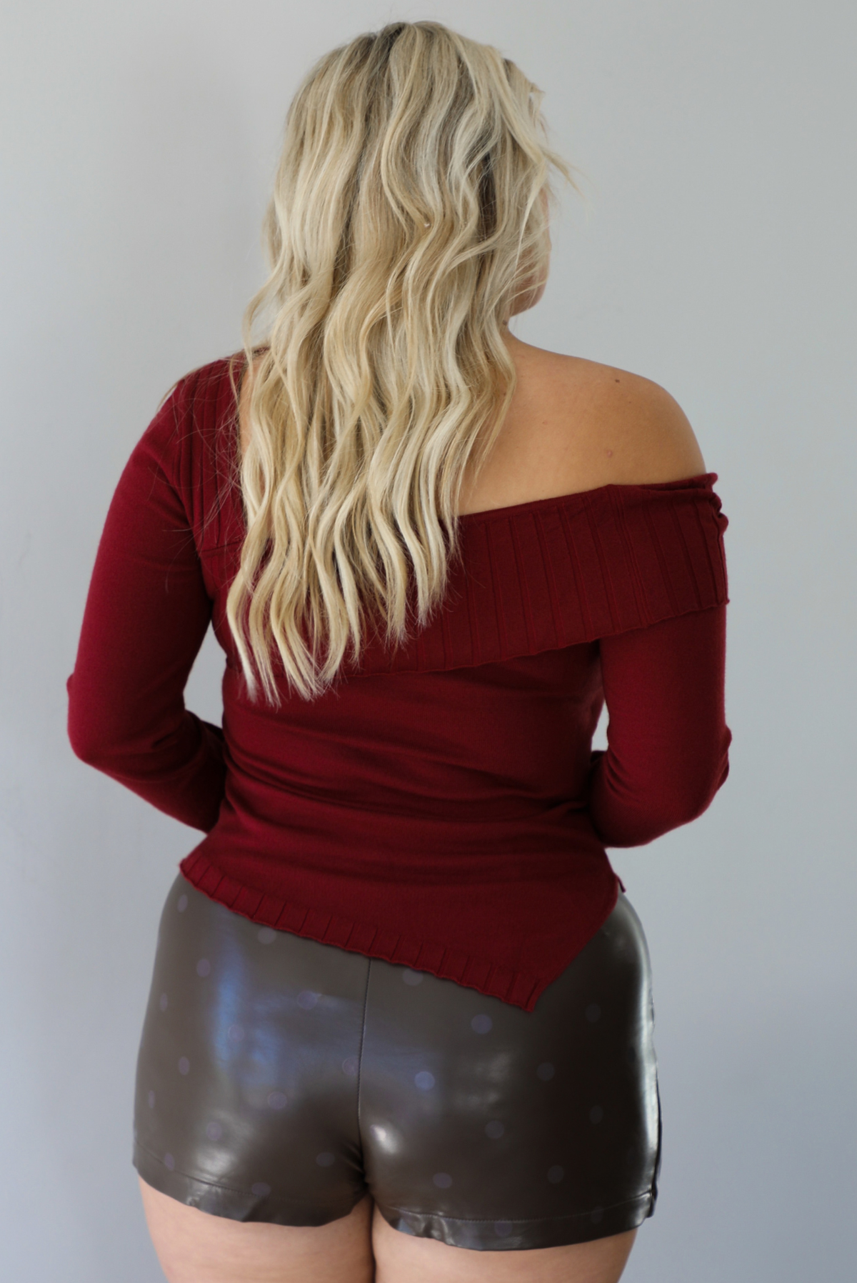 Woman shown from behind wearing a maroon knit, long sleeved, off the shoulder knit top paired with a dark brown vegan leather mini skirt with a monochromatic polka dot print pattern, standing indoors against a white wall.