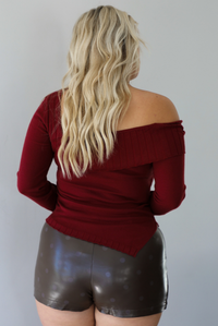 Woman shown from behind wearing a maroon knit, long sleeved, off the shoulder knit top paired with a dark brown vegan leather mini skirt with a monochromatic polka dot print pattern, standing indoors against a white wall.