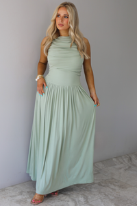 Woman wearing a sage green maxi length dress, which has a rouched detail at the bust, a fitted waistline, a flowy skirt. The photo is taken indoors against a white wall with white marble flooring. 