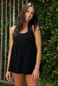 Woman wearing a black romper that looks like a dress, with a halter tie neckline, baby-doll cut, and flowy skirt outdoors with greenery and an iron fence in the background. 