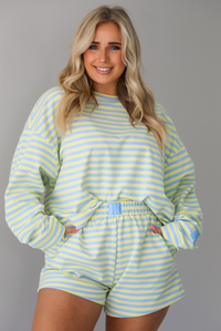 Woman wearing a blue and pale yellow striped two piece set featuring an oversized sweatshirt and shorts. The photo is taken indoors against a white wall and shows a closeup of the set, which is tucked into the front of the shorts. 