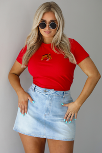 Woman wearing a red fitted baby tee with an embroidered horse graphic on the front. The top is paired with a high waisted, light-wash denim skort. The photo is taken indoors against a white wall.