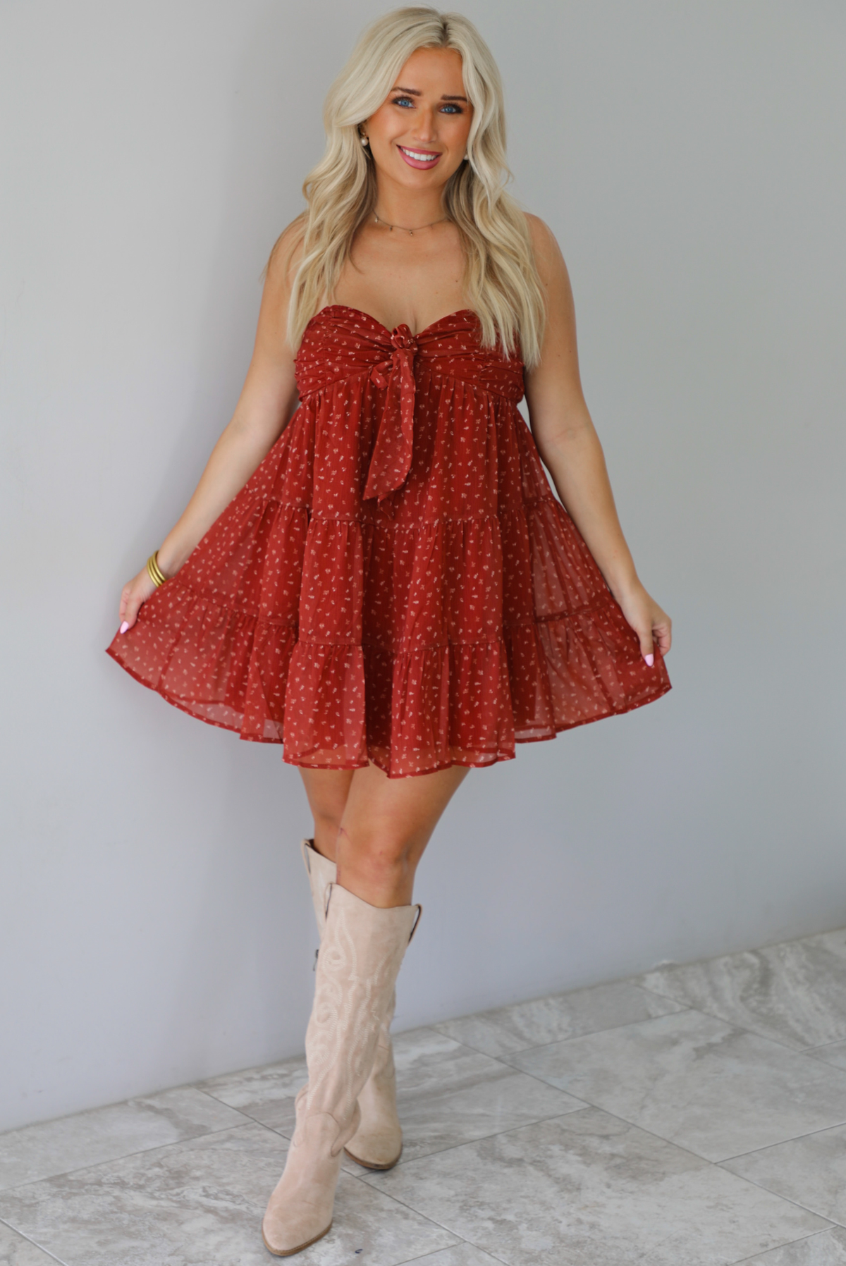 A woman wearing a rust mini length dress with a cream floral and leaf tiny print design throughout, has a strapless design with a tie detail in the center of the bust, with a baby-doll dress cut that's fitted at the bust with a flowy skirt, standing indoors against a white wall with white marble flooring. 
