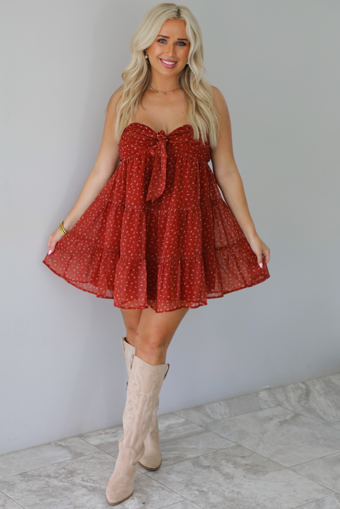 A woman wearing a rust mini length dress with a cream floral and leaf tiny print design throughout, has a strapless design with a tie detail in the center of the bust, with a baby-doll dress cut that's fitted at the bust with a flowy skirt, standing indoors against a white wall with white marble flooring. 