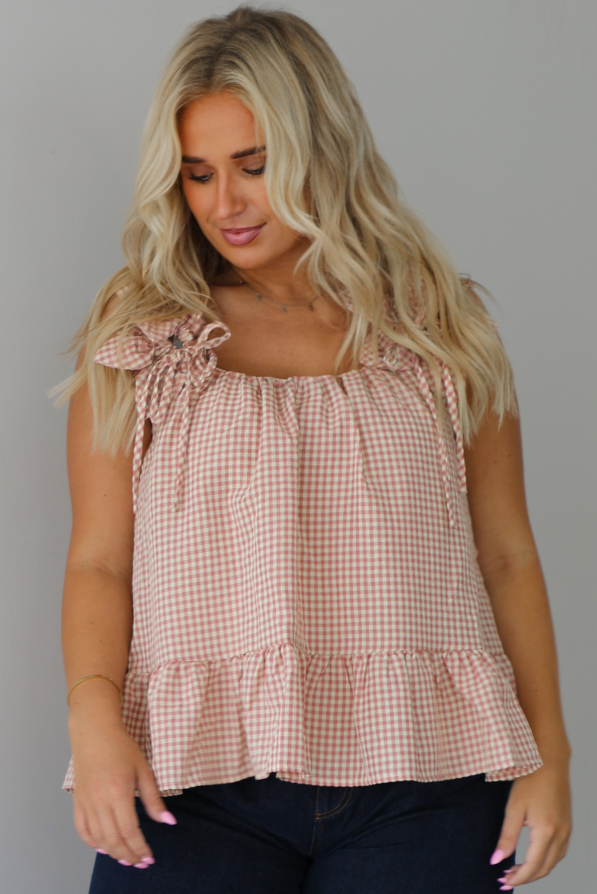 A woman wearing a light maroon and cream gingham flowy top with dark denim jeans, standing indoors against a white wall.
