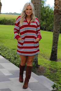 A woman wearing a red burgundy mini dress with a white and blue striped pattern throughout, collared neckline, long sleeves, and functional pockets on ether side, standing outdoors on a stone path with palm trees and other greenery in the background. 