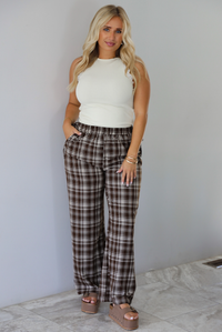 A woman modeling a pair of brown and sand plaid wide-leg pants with a cream ribbed sleeveless top, standing indoors against a white wall with white marble flooring. 
