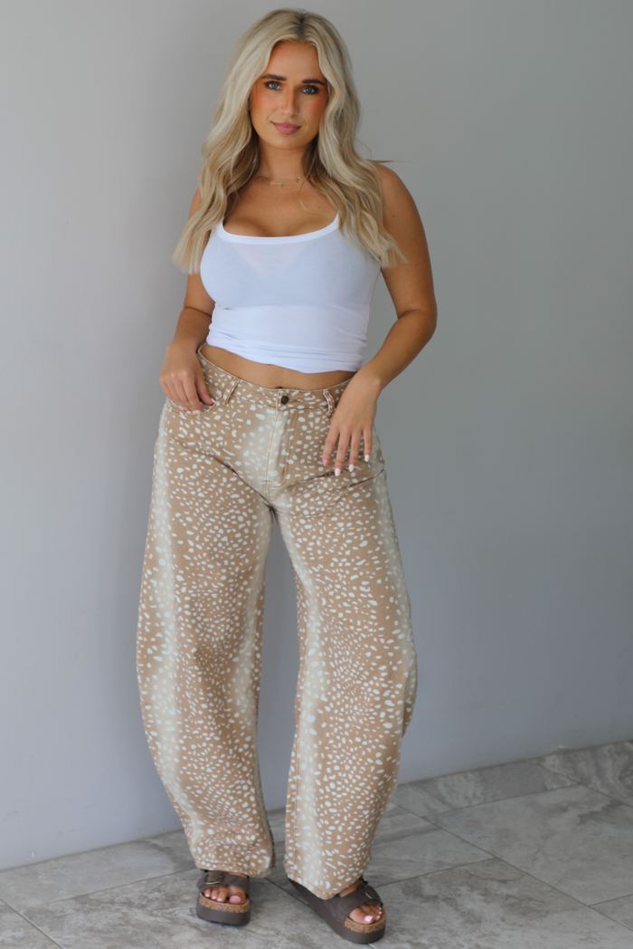 Woman wearing a white, ribbed, cropped tank with mid-rise tan and sand barrel denim with a doe pattern indoors against a white wall with white marble flooring. 