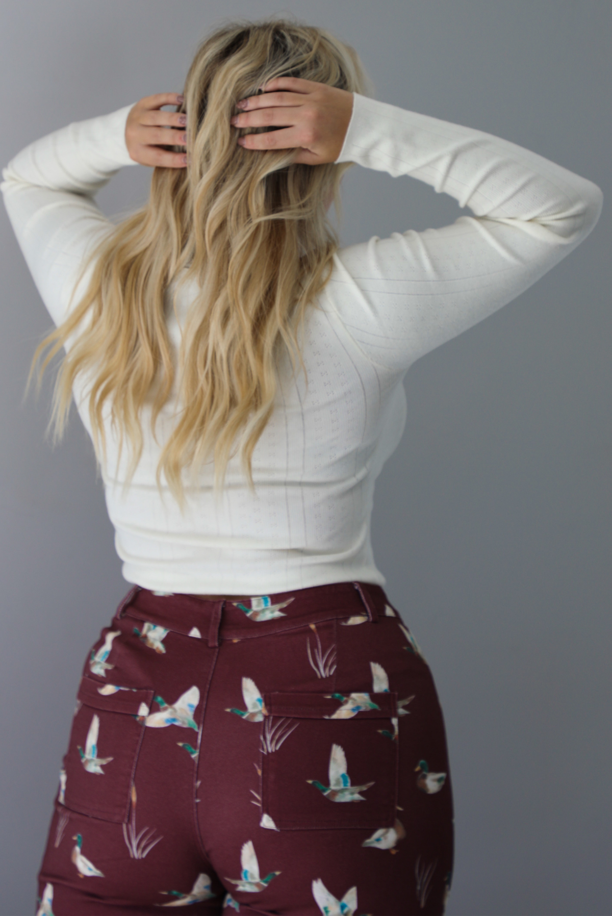 A woman shown from behind wearing a cream colored knit long sleeved top paired with high waisted maroon jeans with a repeating mallard design, standing indoors against a white wall.