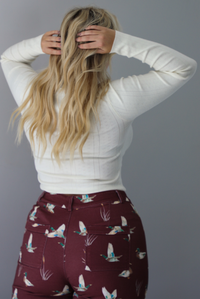 A woman shown from behind wearing a cream colored knit long sleeved top paired with high waisted maroon jeans with a repeating mallard design, standing indoors against a white wall.
