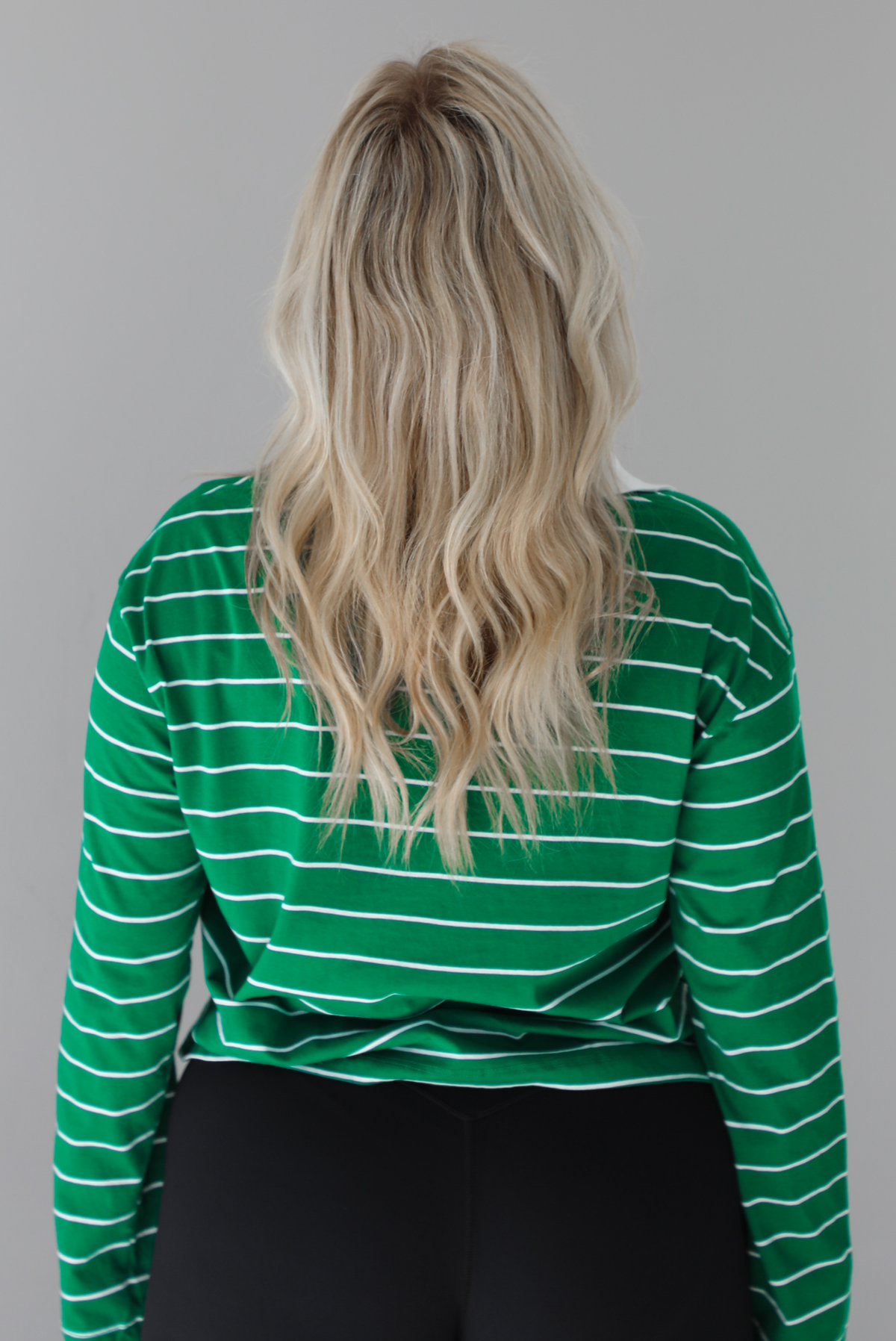 Woman wearing a green with white striped collared cropped top with functional button closures a quarter at the top. The top is paired with black leggings. The photo shows the back of the top. The photo is taken in front of a white wall.
