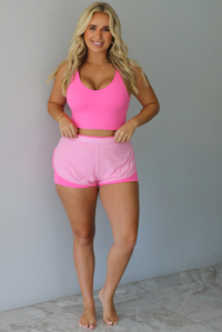 A woman modeling a bright pink two-piece activewear set consisting of a scoop-neck crop top and matching athletic shorts with a darker, hot pink trim, standing indoors against a white wall with white marble flooring. 