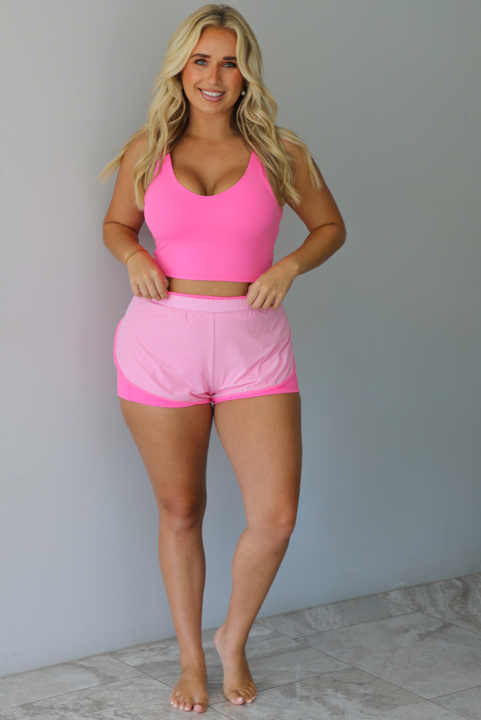 A woman modeling a bright pink two-piece activewear set consisting of a scoop-neck crop top and matching athletic shorts with a darker, hot pink trim, standing indoors against a white wall with white marble flooring. 