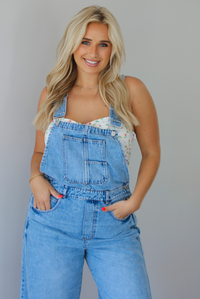 Woman wearing a pair of denim overalls with a white floral strapless top underneath indoors against a white wall.
