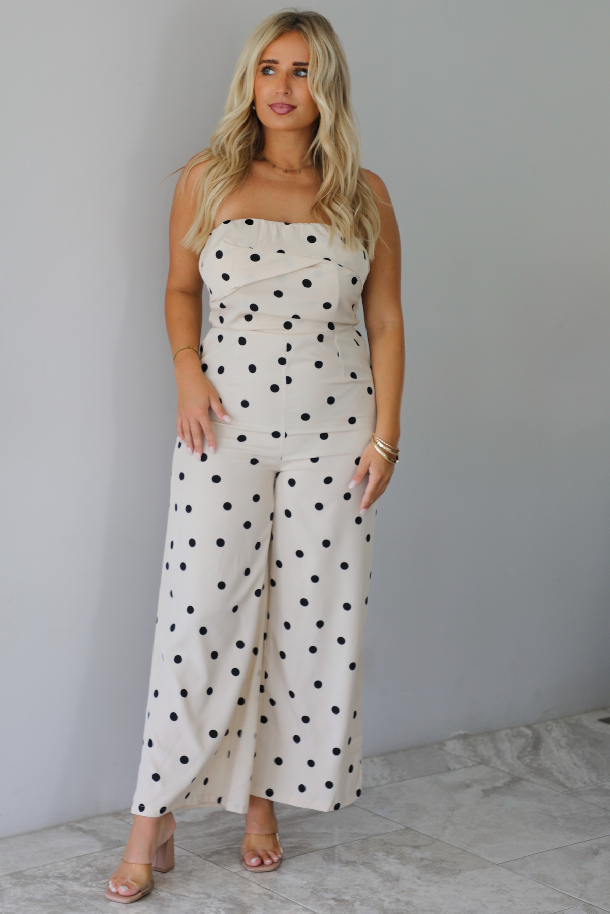 A woman wearing a sand strapless jumpsuit with a black polka dot print pattern throughout, standing indoors against a white wall with white marble flooring. 