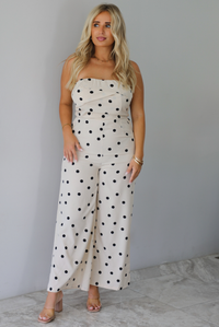 A woman wearing a sand strapless jumpsuit with a black polka dot print pattern throughout, standing indoors against a white wall with white marble flooring. 