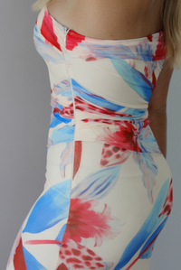 A woman modeling a strapless off-the-shoulder maxi dress with a vibrant red, blue, and cream floral print against a white wall. The photo shows a closeup of the side of the dress, which has a zipper closure. 