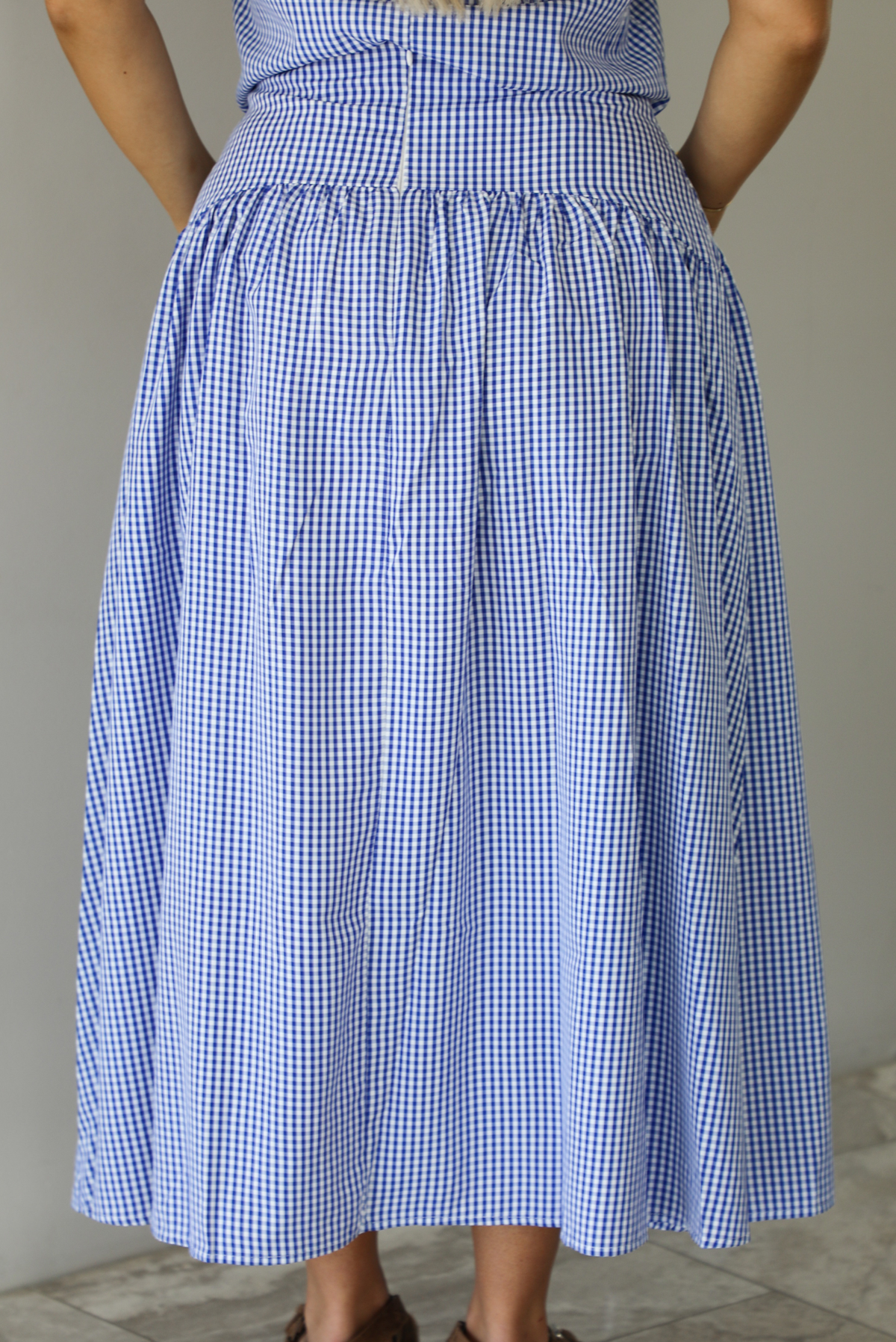 Woman wearing a blue and white gingham two piece set featuring a sleeveless top and high waisted, maxi length skirt indoors against a white wall with white marble flooring. The photo shows a closeup of the back of the skirt, which has a zipper closure in the center. 