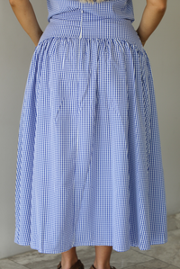 Woman wearing a blue and white gingham two piece set featuring a sleeveless top and high waisted, maxi length skirt indoors against a white wall with white marble flooring. The photo shows a closeup of the back of the skirt, which has a zipper closure in the center. 