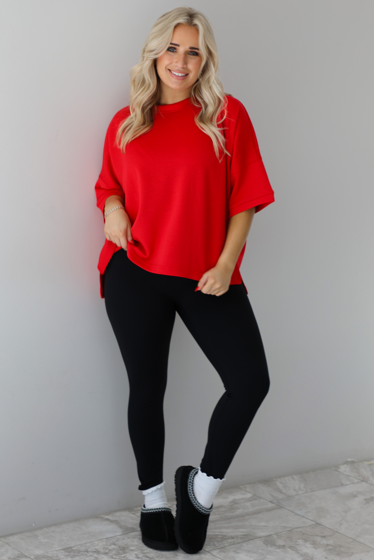 Woman wearing an oversized red t-shirt with black, fitted lettings, scrunchie socks, and black ugg boots. The photo is taken indoors against a white wall with white marbled flooring. 