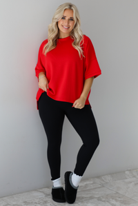 Woman wearing an oversized red t-shirt with black, fitted lettings, scrunchie socks, and black ugg boots. The photo is taken indoors against a white wall with white marbled flooring. 