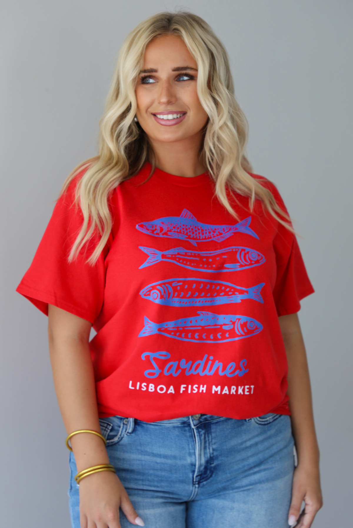 A woman wearing a red graphic t-shirt with blue sardine illustrations and the text 'Sardines LISBON FISH MARKET', paired with blue jeans, standing indoors against a white wall.
