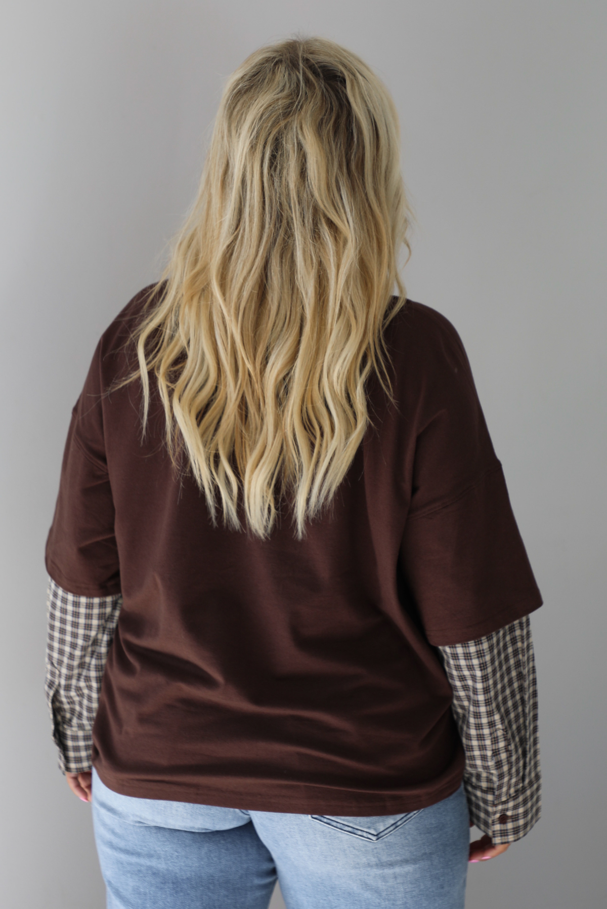 A woman shown from behind wearing a brown t-shirt with a layered look of plaid sleeve detail, paired with light wash denim jeans, standing indoors against a white wall.