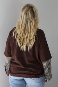 A woman shown from behind wearing a brown t-shirt with a layered look of plaid sleeve detail, paired with light wash denim jeans, standing indoors against a white wall.